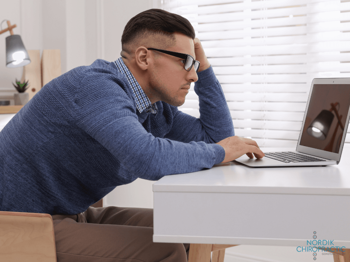Person in glasses leaning toward a laptop at a desk, looking tired in a bright home office.