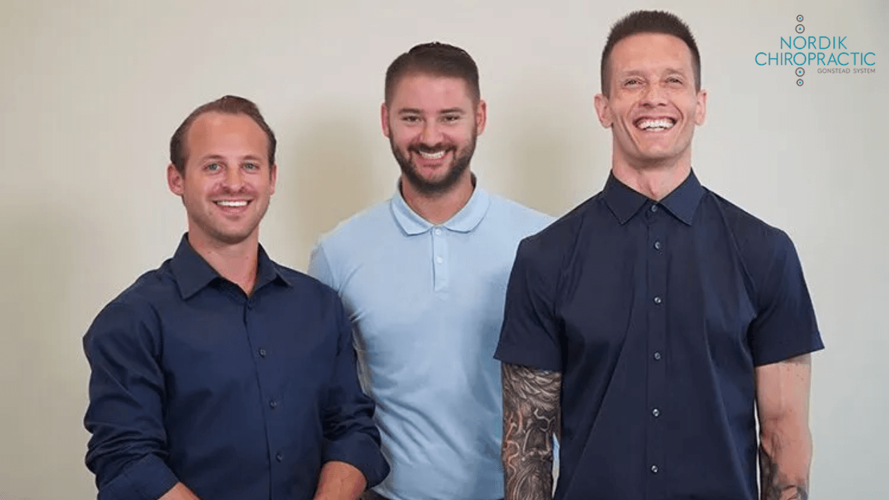 Three smiling people standing against a light wall, wearing navy and light blue shirts.