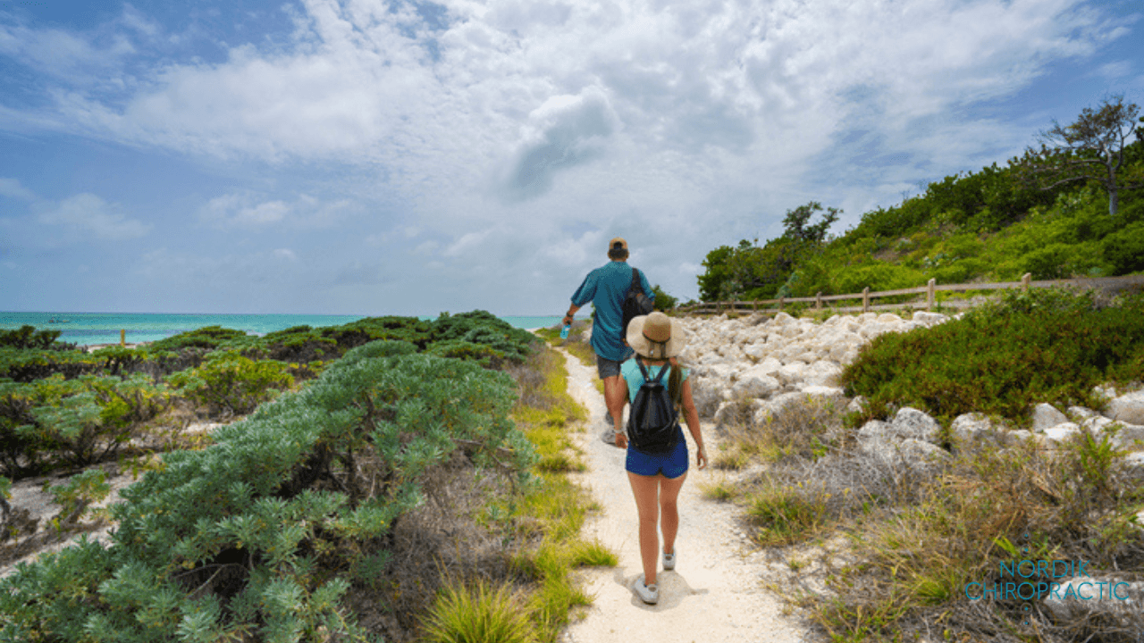 Two people walking a sandy trail beside rocky green dunes with ocean and cloudy sky in view