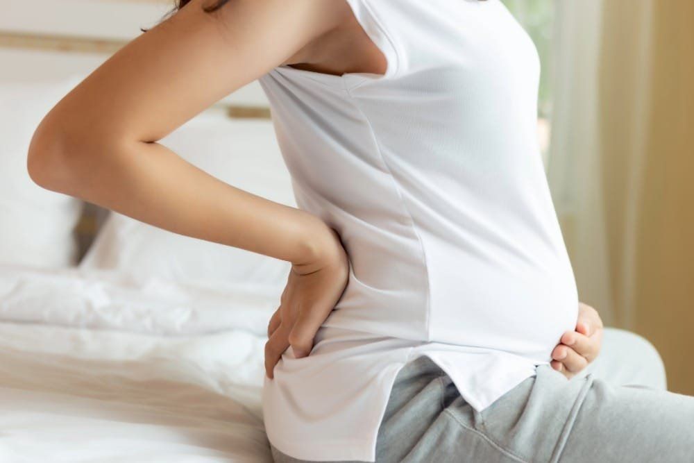 Person sitting on bed, holding lower back in pain, wearing white shirt and gray pants