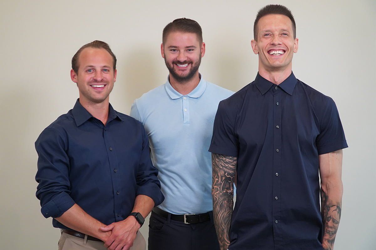 Three smiling men stand side by side indoors against a plain background, wearing button-up shirts.