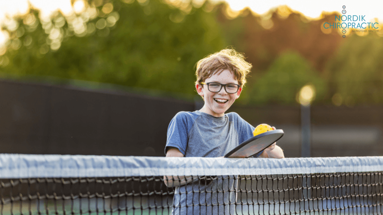 Smiling child with glasses holding a tennis racket by the court net at sunset