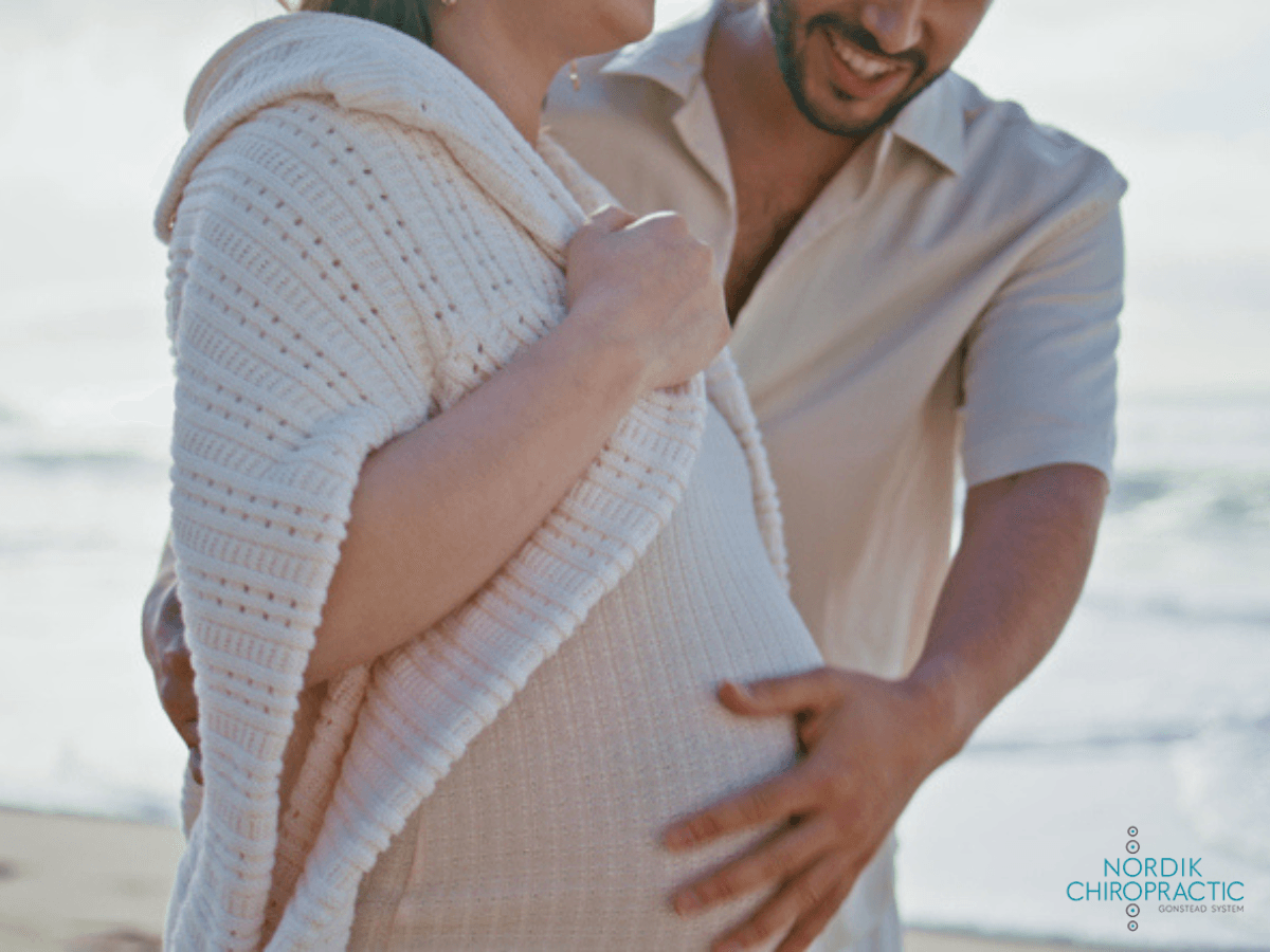 Couple embracing on a beach, with the pregnant woman wrapped in a white knit shawl at sunset