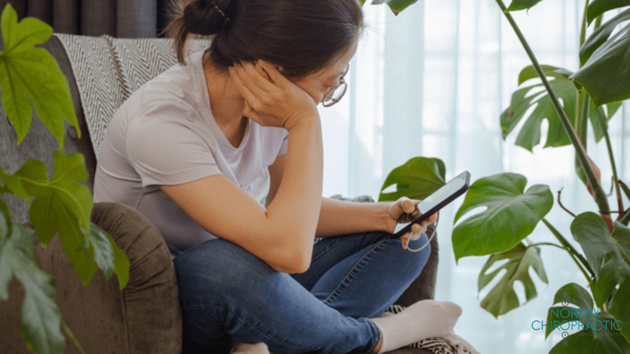 Person sitting on a couch, holding head in hand while looking at a phone, in a bright room with houseplants.