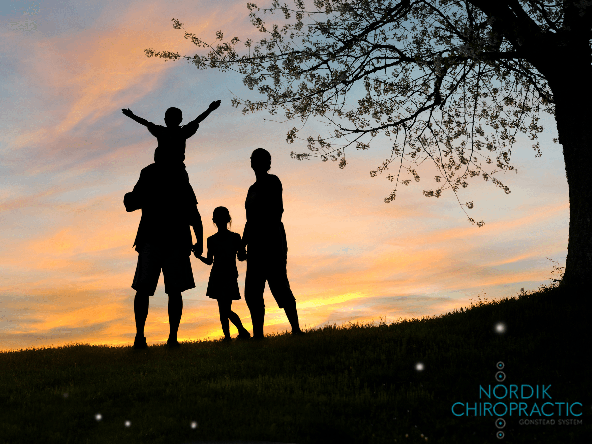 Silhouetted family standing at sunset under a tree, one child on shoulders, arms raised