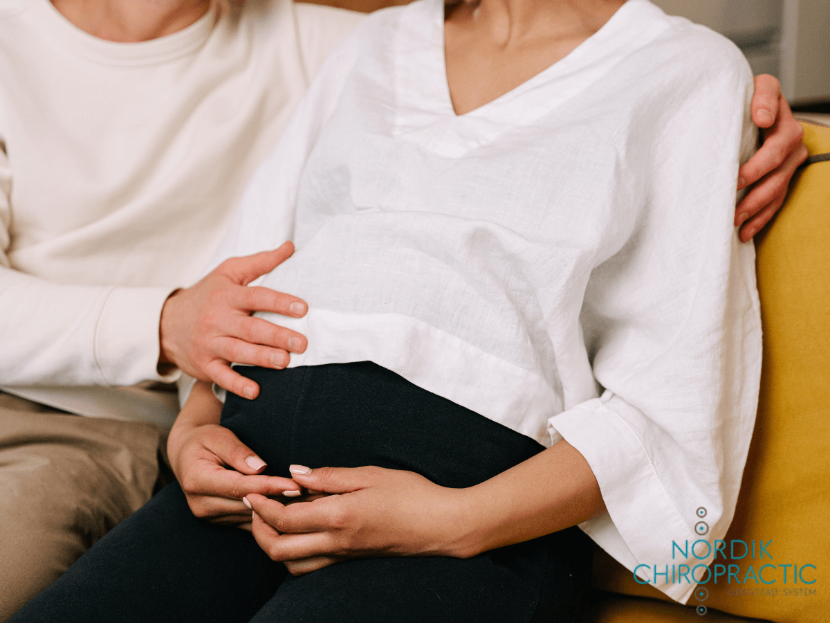 Two people sit close together, hands gently cradling a pregnant belly in a white shirt.