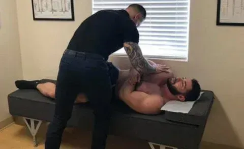Two men on a massage table; one applies pressure to the other's upper body in a clinic room.