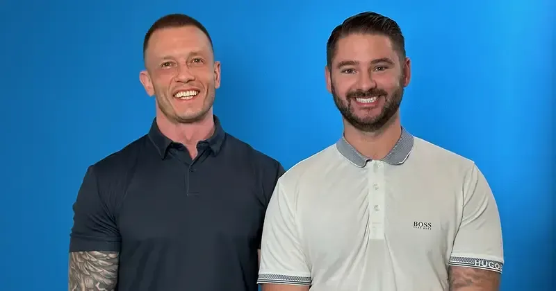 Two smiling men standing side by side against a blue background, wearing collared shirts.