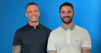 Two smiling men standing side by side against a blue background, wearing collared shirts.