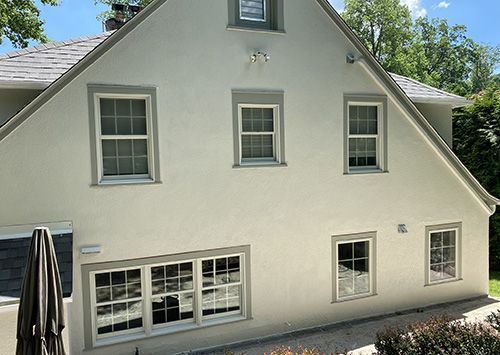 Exterior view of a light-colored stucco house with gray trim, windows, and a gray roof on a sunny day.