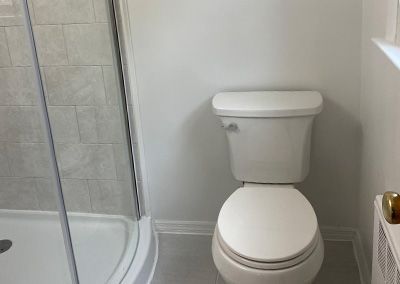 White toilet in a bathroom, next to a shower with a glass door. Walls are white and light gray tile.