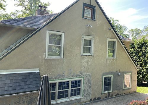 Back of a two-story beige stucco house with several windows, dark roof, and a chimney.