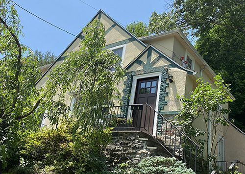 Two-story house with tan stucco, green trim, and dark brown door, approached by stone steps, surrounded by greenery.