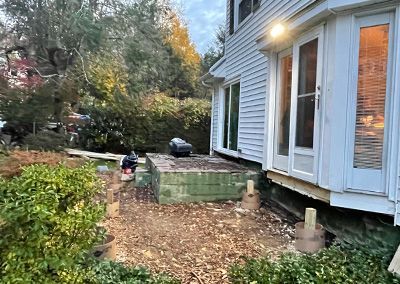 Backyard scene: a house with a door, a wooden deck under construction, and foliage.