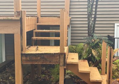 Wooden deck and stairs with visible damage, set against a building and greenery.