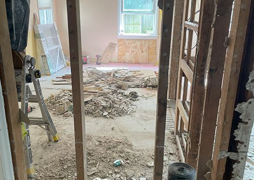 Room under renovation, debris on floor, exposed framing. Window visible in the background.