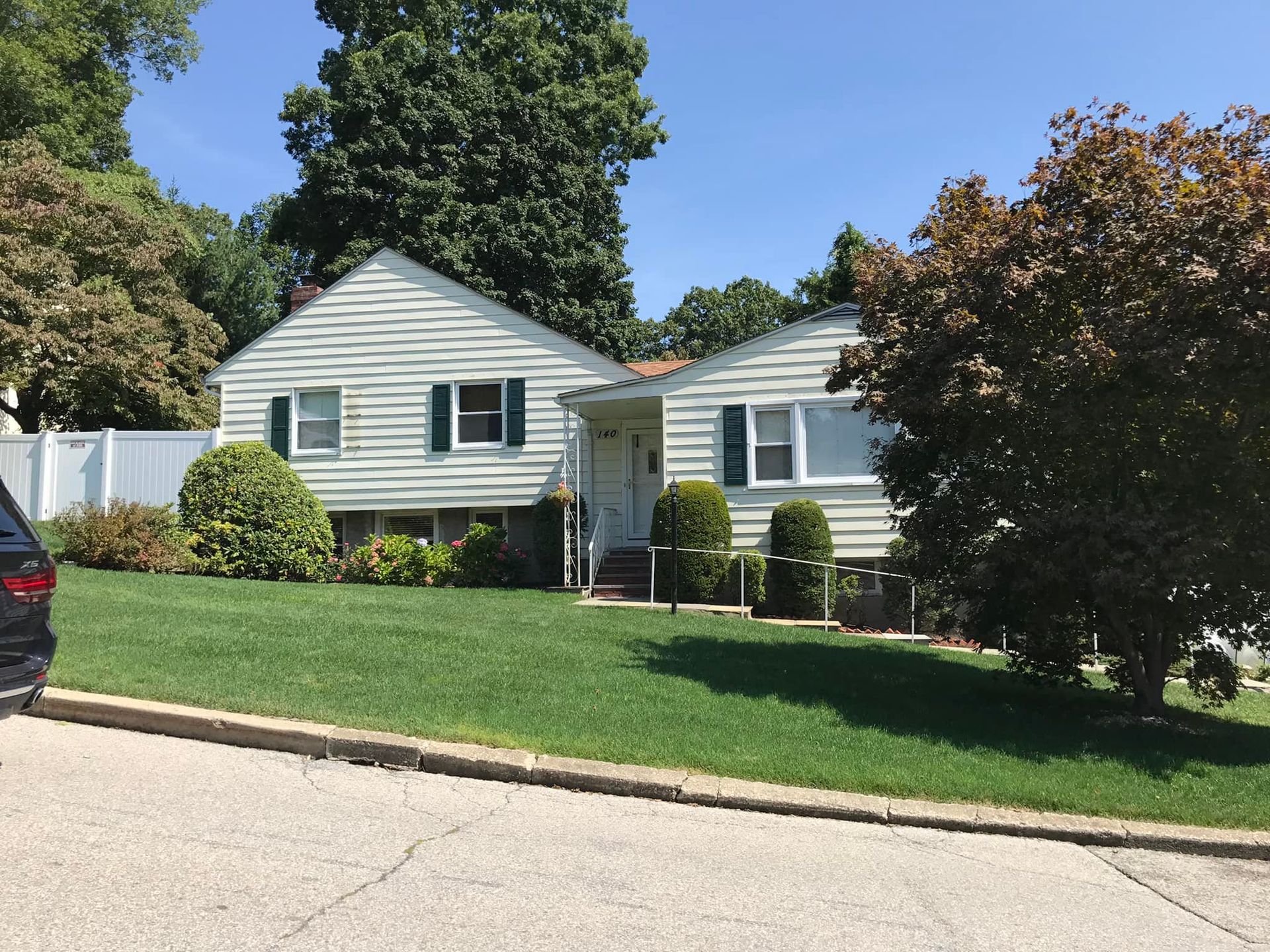 Beige house with green shutters, lawn, and surrounding trees on a sunny day.