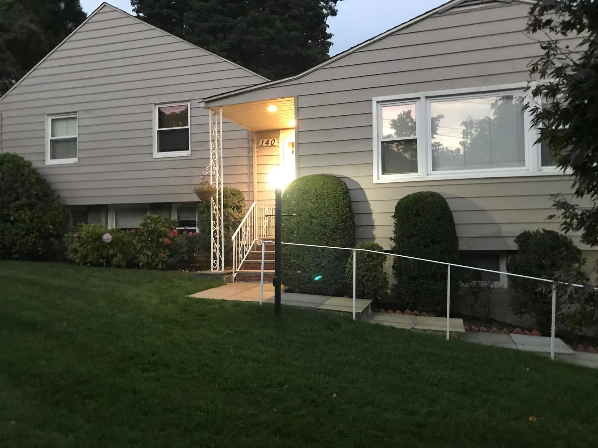 Two-story gray house with a small front porch, lit with a light. A green lawn slopes down from the house.