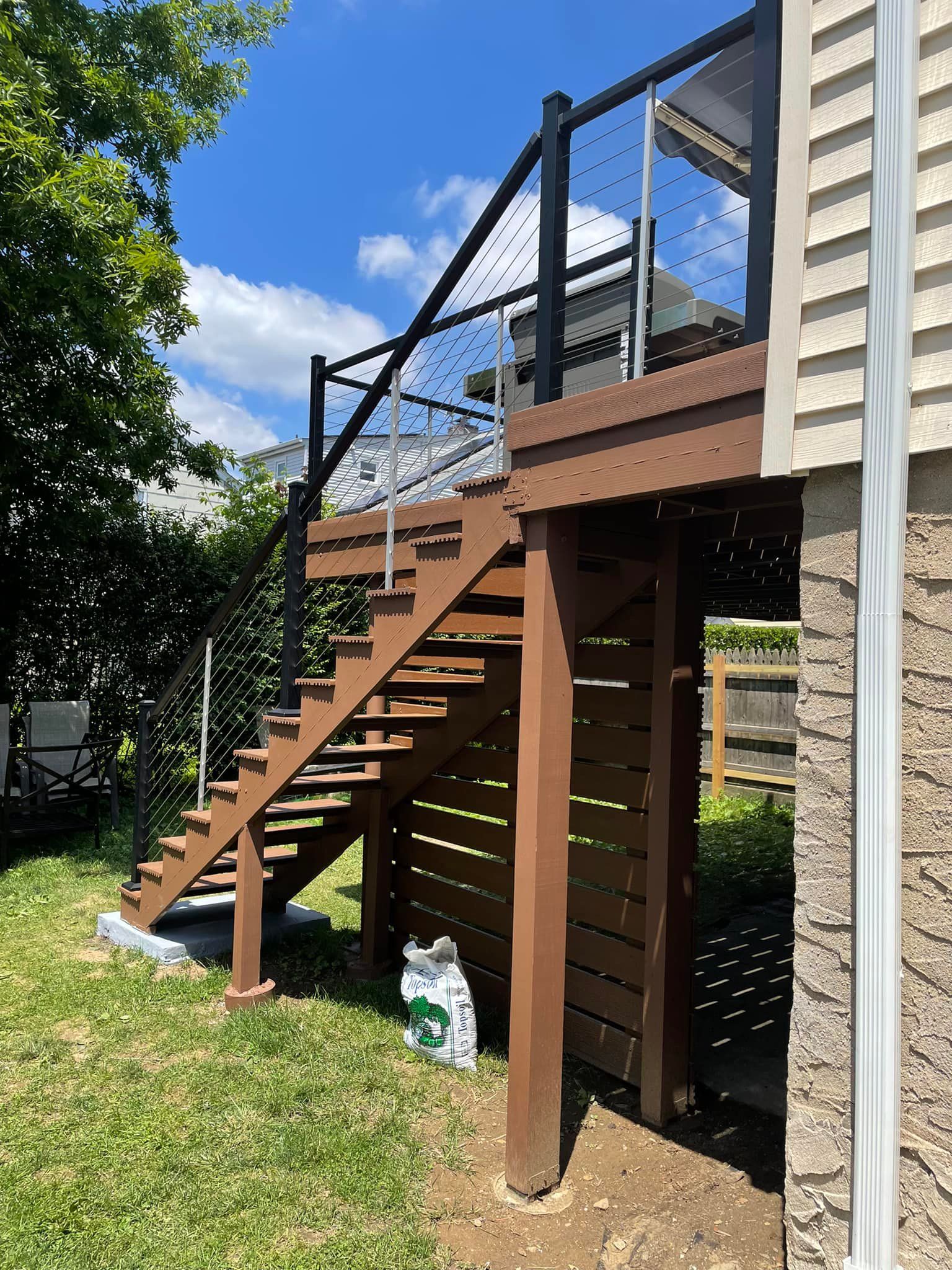 Brown wooden deck and stairs with black railing, next to a house on a sunny day.