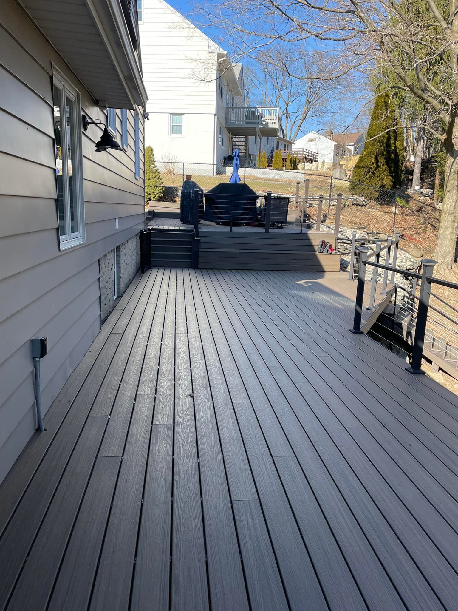 Wooden deck beside a house with a grill and stairs leading to a yard, on a sunny day.