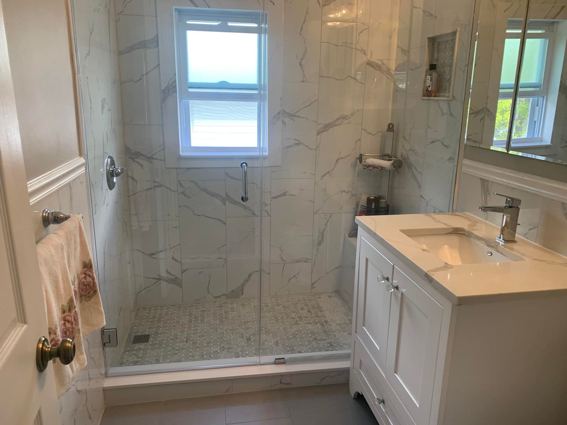 Bathroom with white marble tile, glass shower, white vanity, and small window.