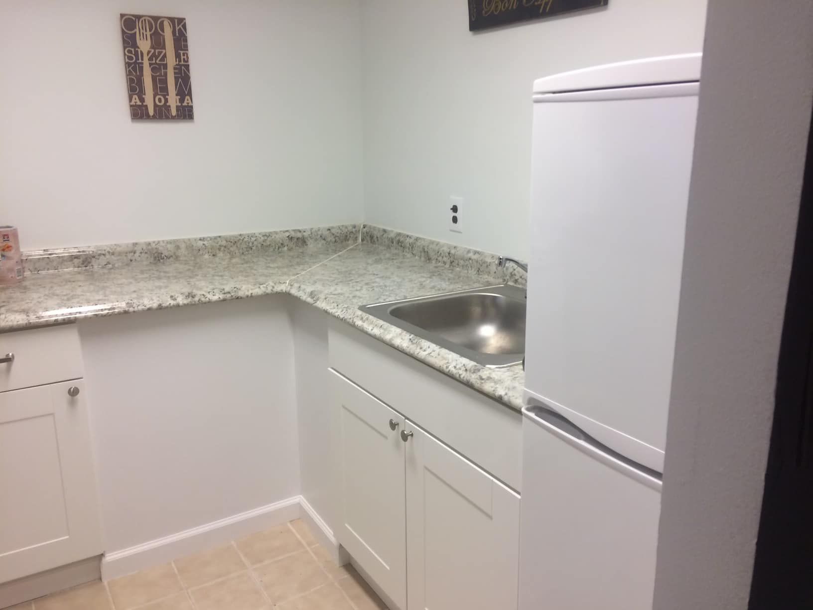 White kitchenette with granite countertop, stainless steel sink, and refrigerator.