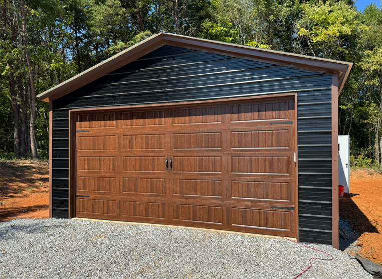 A detached garage with black metal siding and a brown faux-wood carriage-style door, set on a gravel lot near woods.