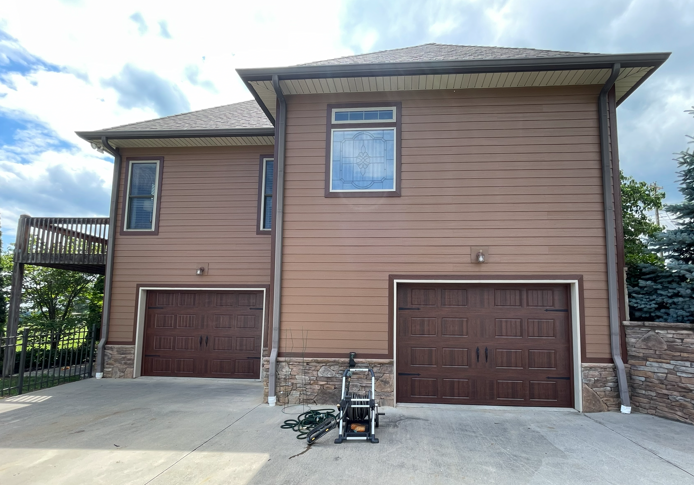 A two-story house with brown siding and stone base, featuring two garage doors and a deck, under a partly cloudy sky.