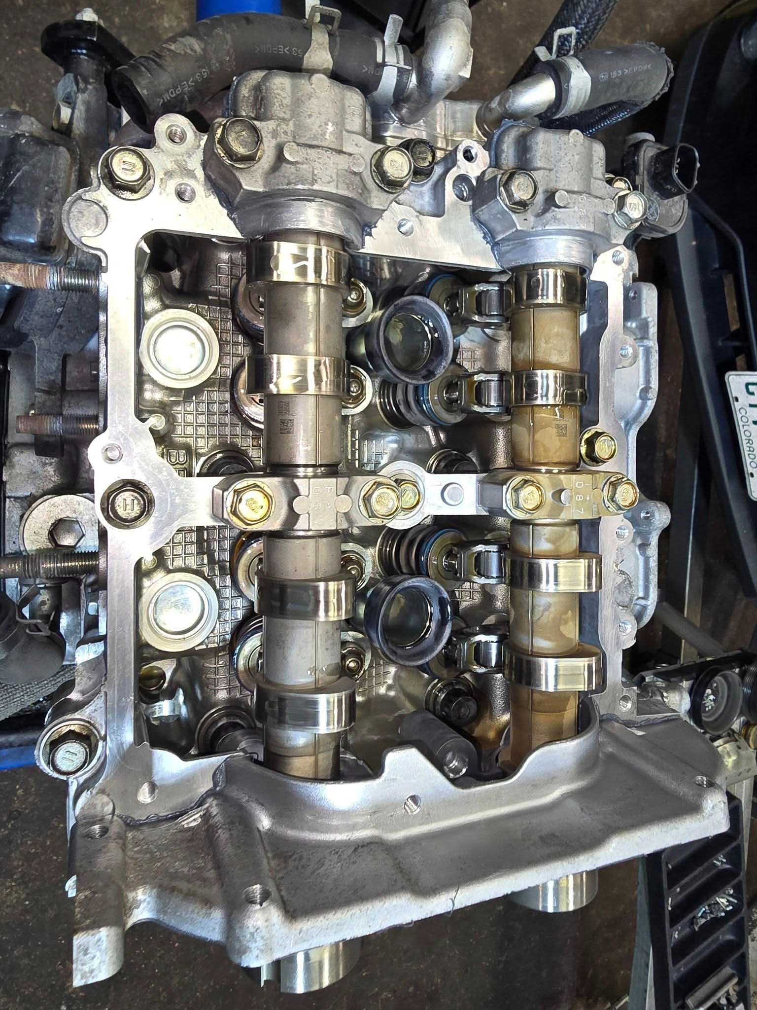 Engine cylinder head with camshafts and valves exposed, viewed from above. Metal and mechanical parts.