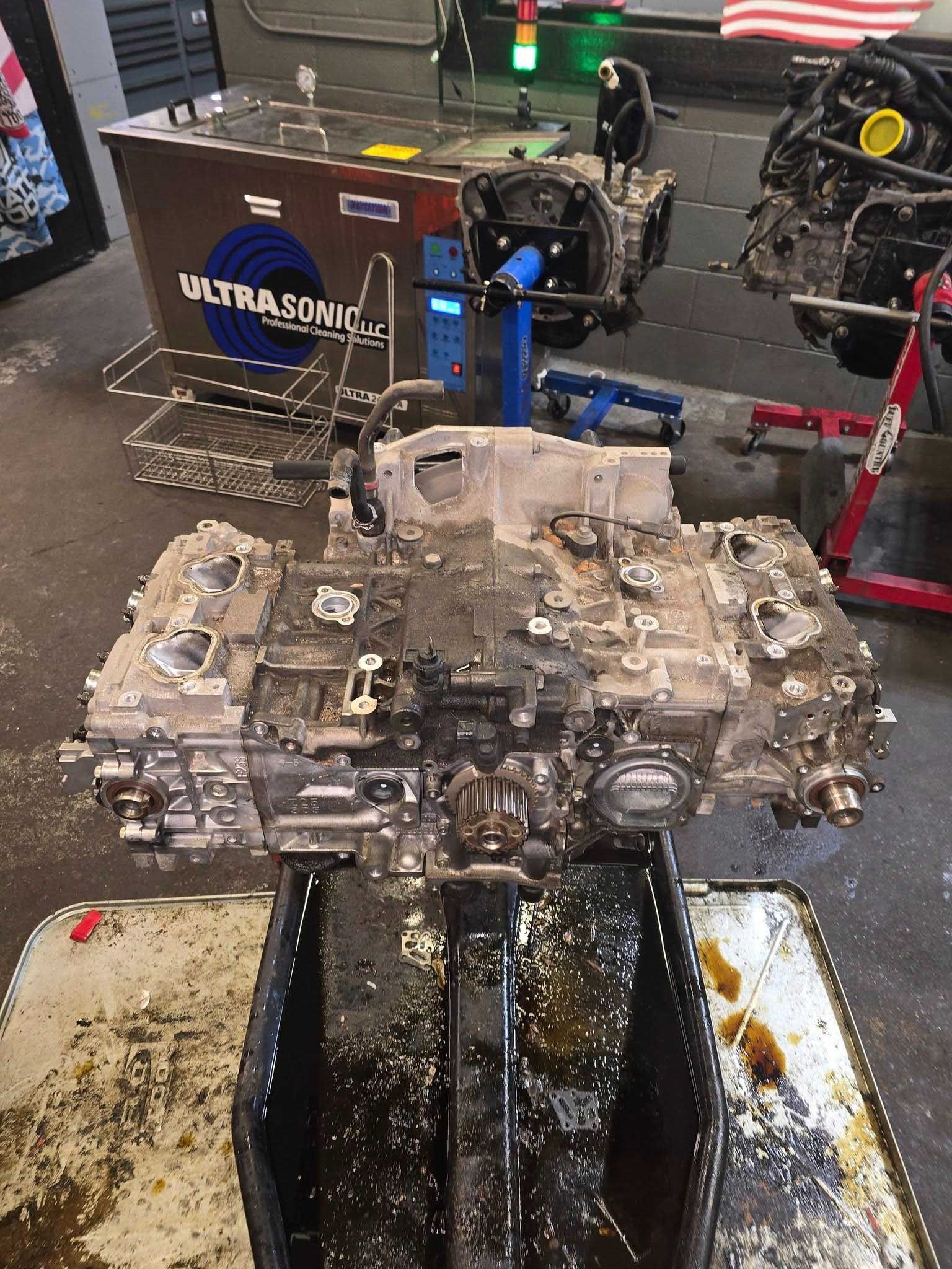 Dirty engine block over a drain pan in a shop, with an ultrasonic cleaner in the background.