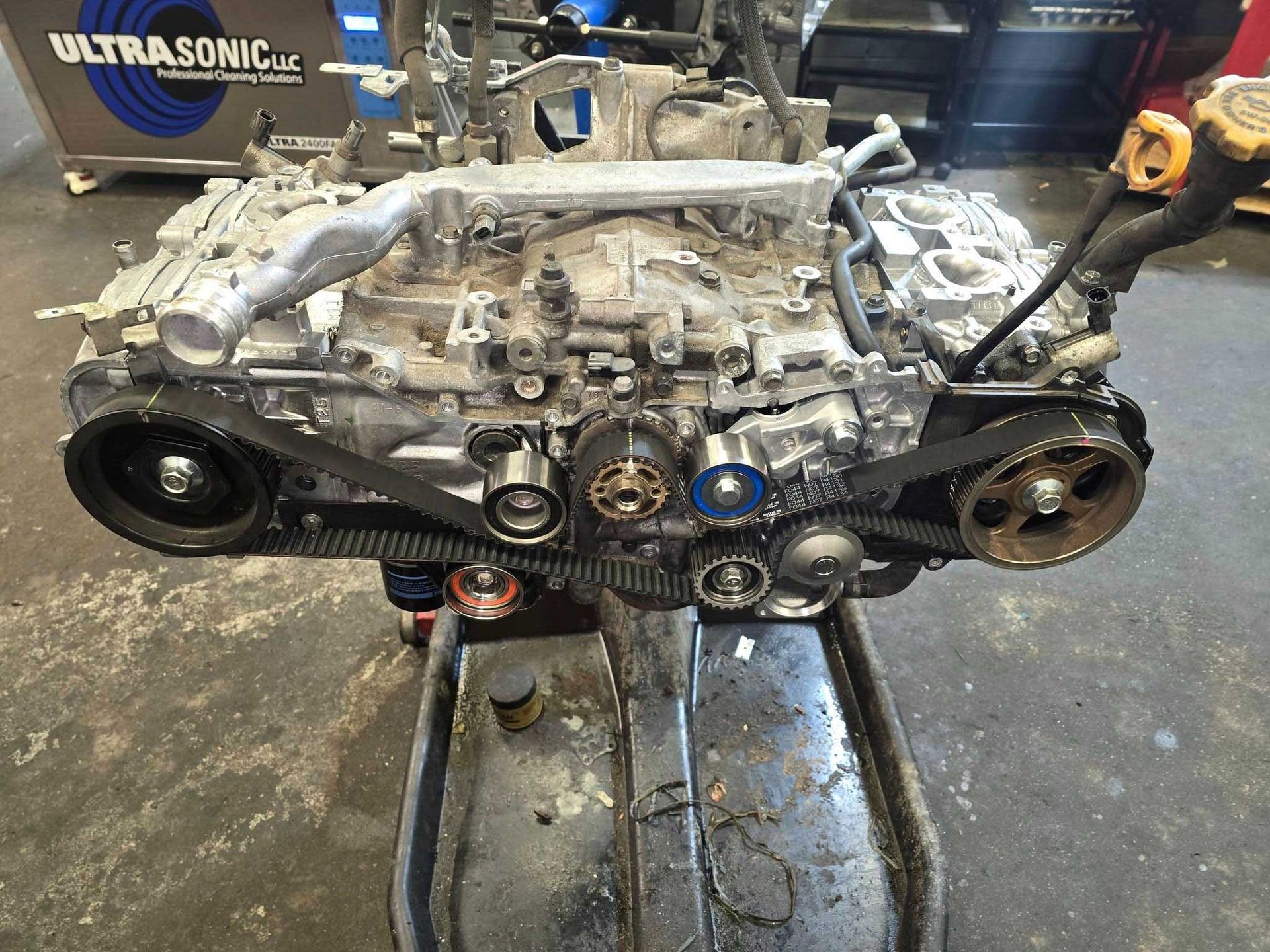 Subaru engine with visible timing belt and pulleys, in a mechanic's shop, mounted above an oil pan.