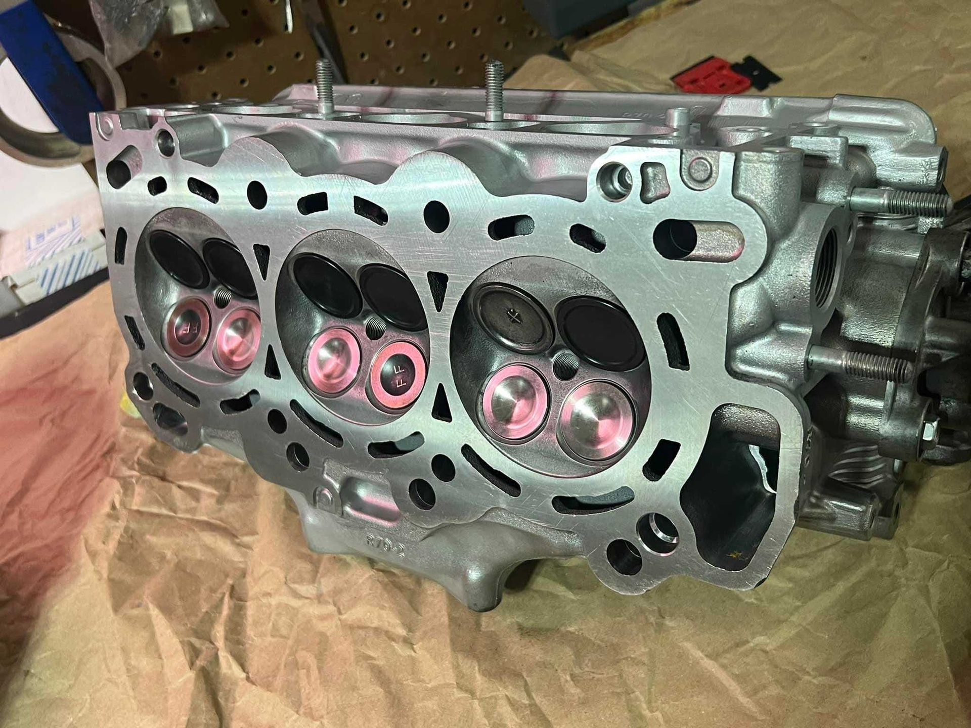Engine cylinder head with valves visible. Silver metal with pink valve stems, resting on brown paper.