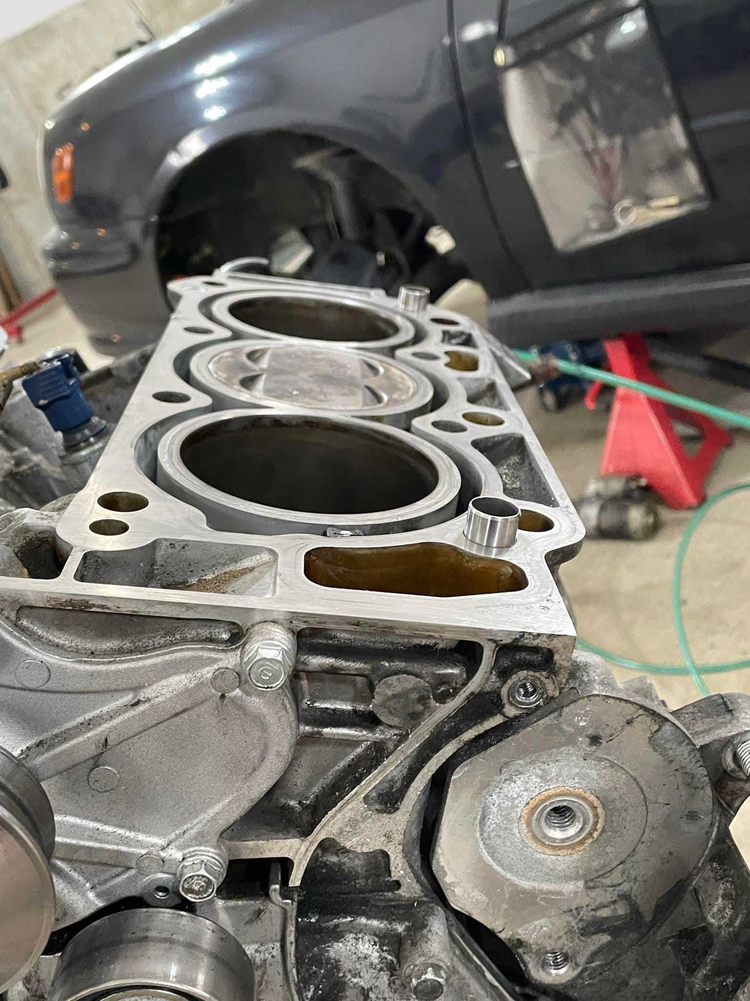 Engine block with cylinder head removed, revealing cylinders. A dark car is in the background.