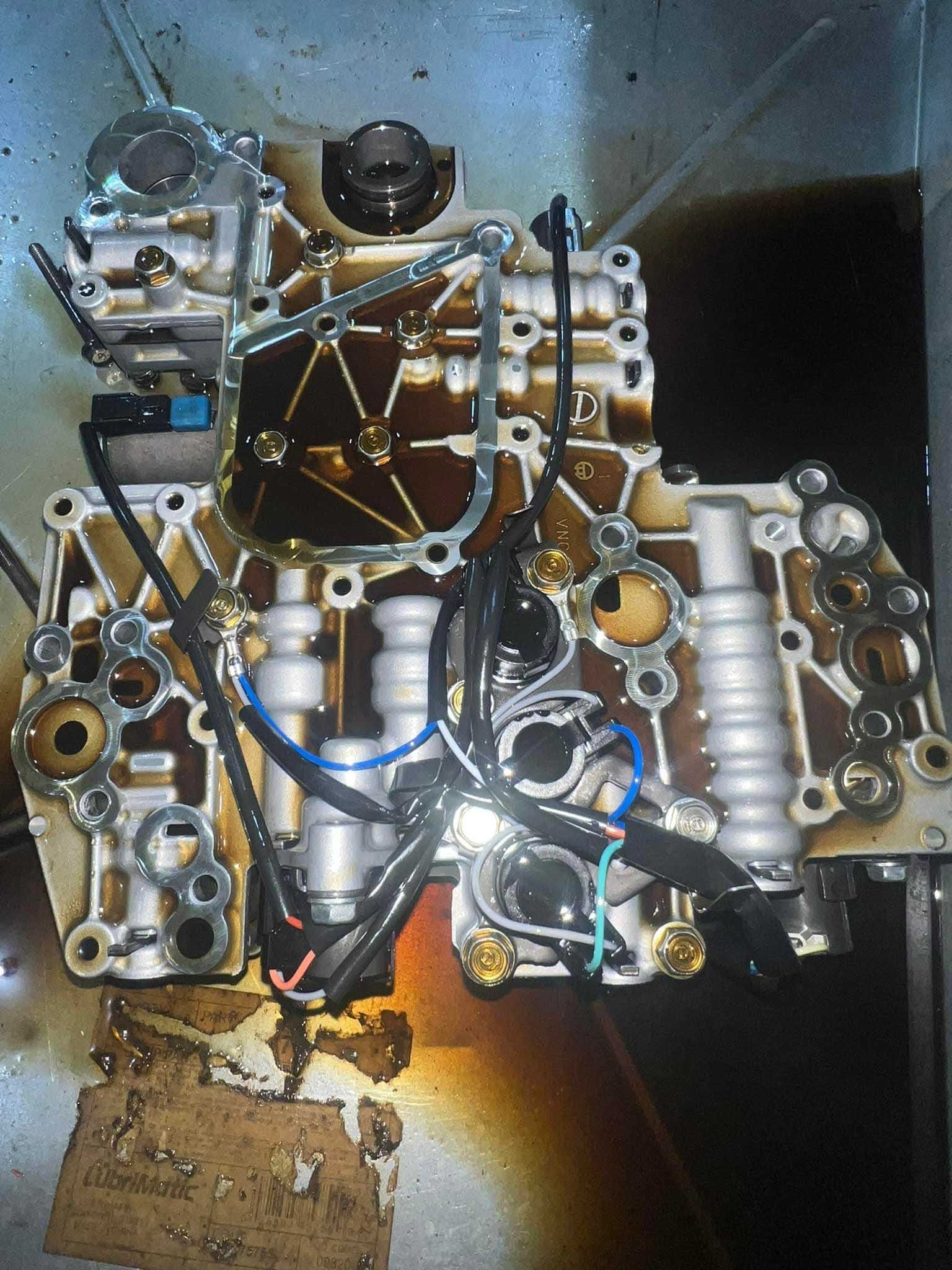 Automatic transmission valve body, open and coated with dark fluid, wiring visible.