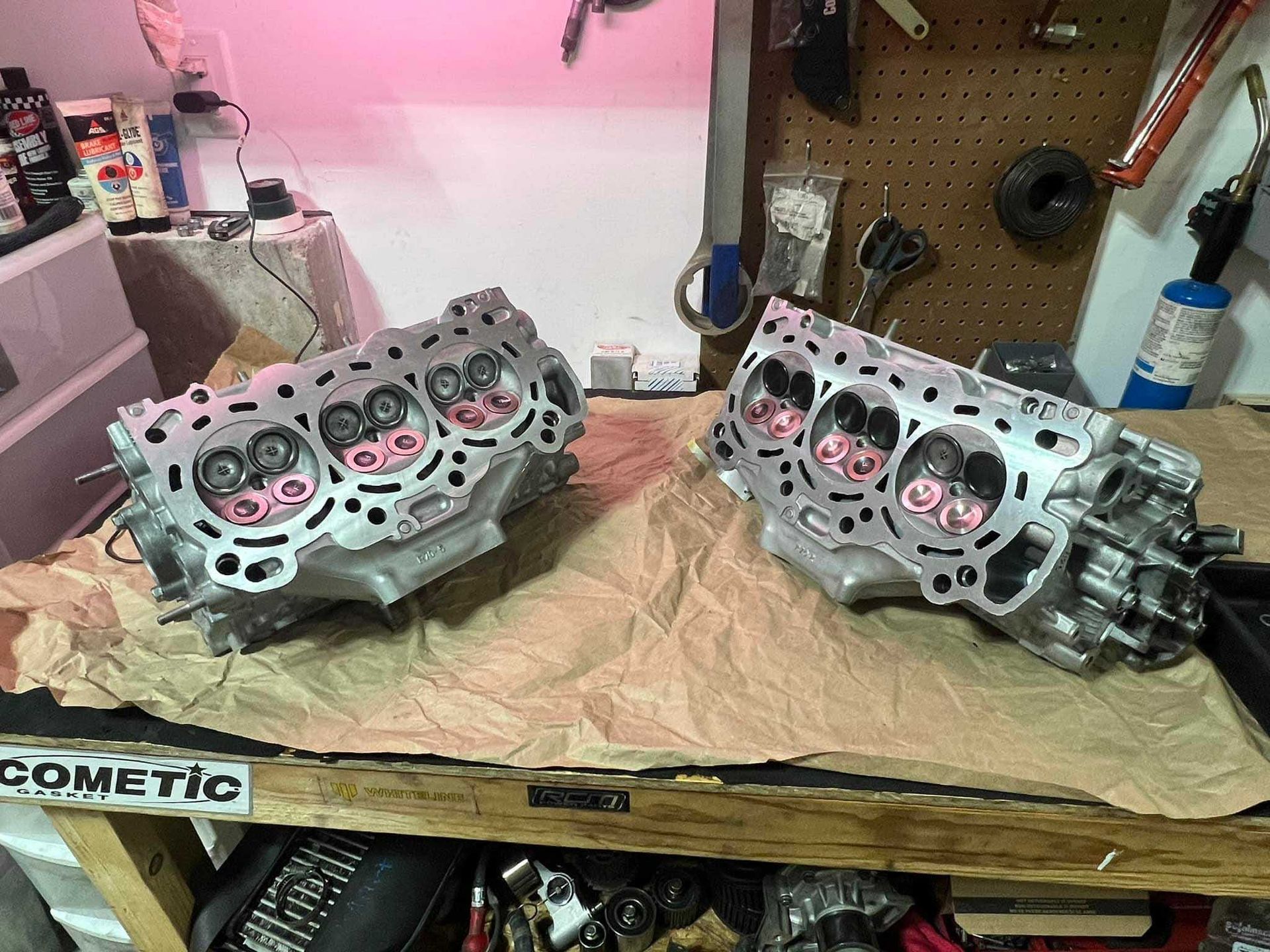 Two engine cylinder heads on a workbench, valves visible. Workshop setting with tools and supplies.