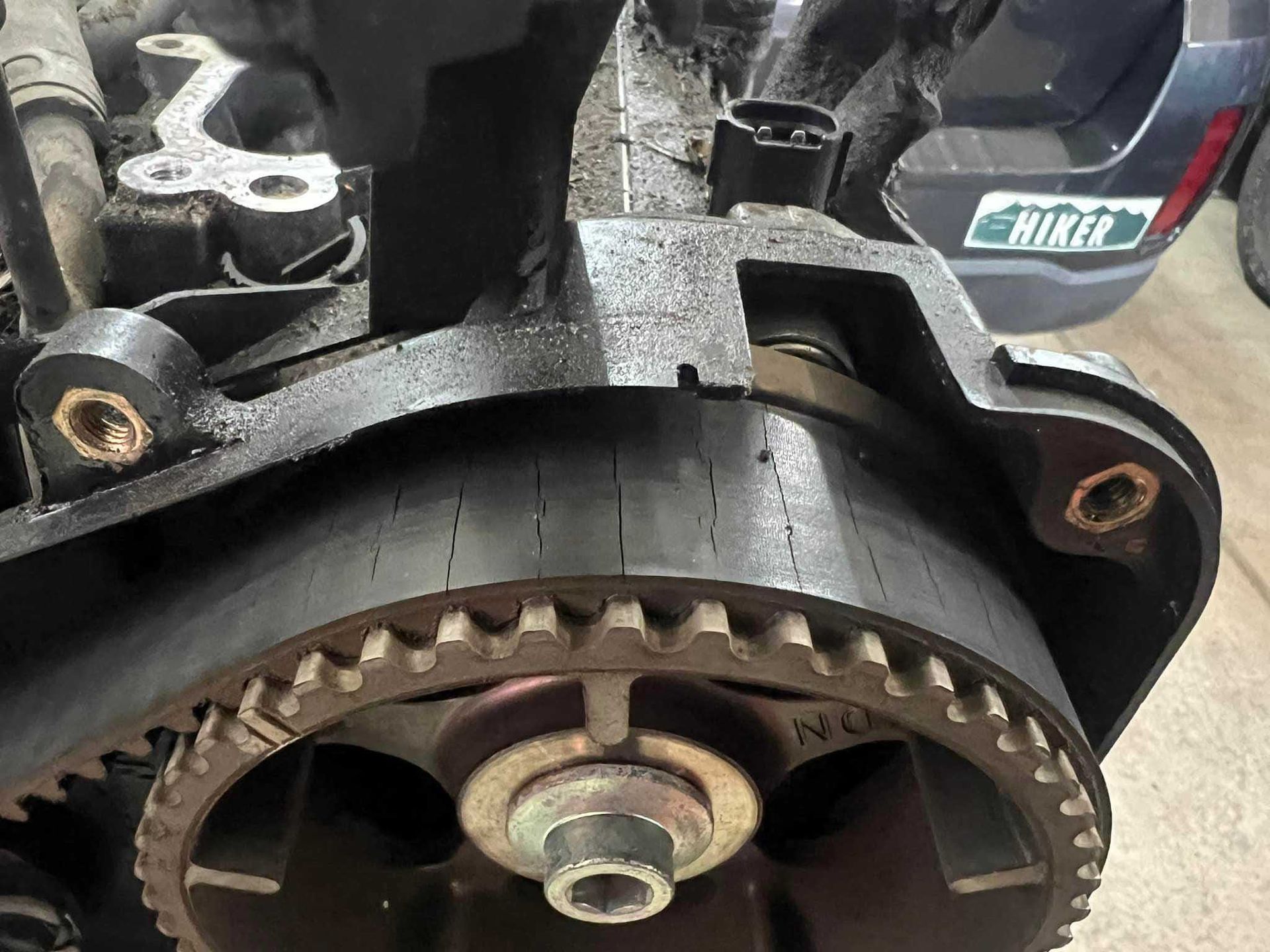 Close-up of a cracked timing belt on an engine. Dark metal and belt material, with visible wear.