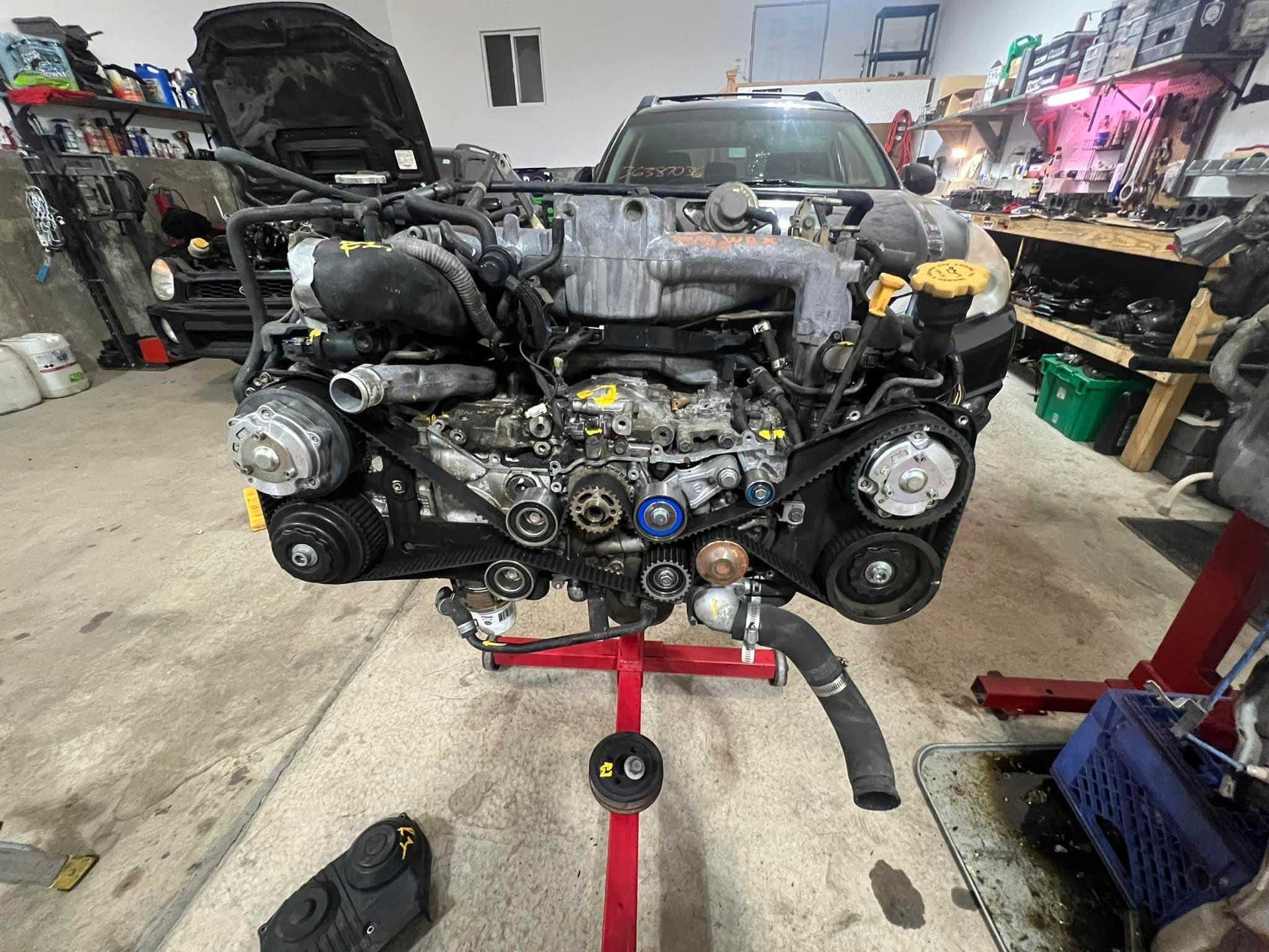 Subaru engine mounted on a red stand, partially disassembled. Garage setting with car in the background.