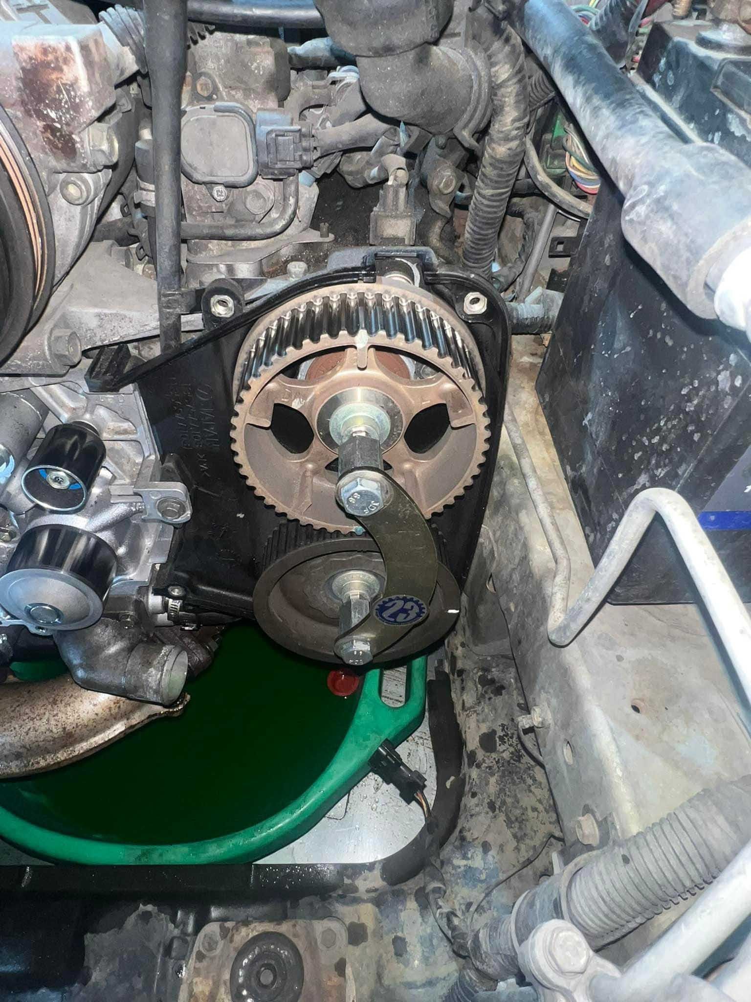 Engine with exposed timing belt and pulleys during repair; oily, green fluid in a catch pan.