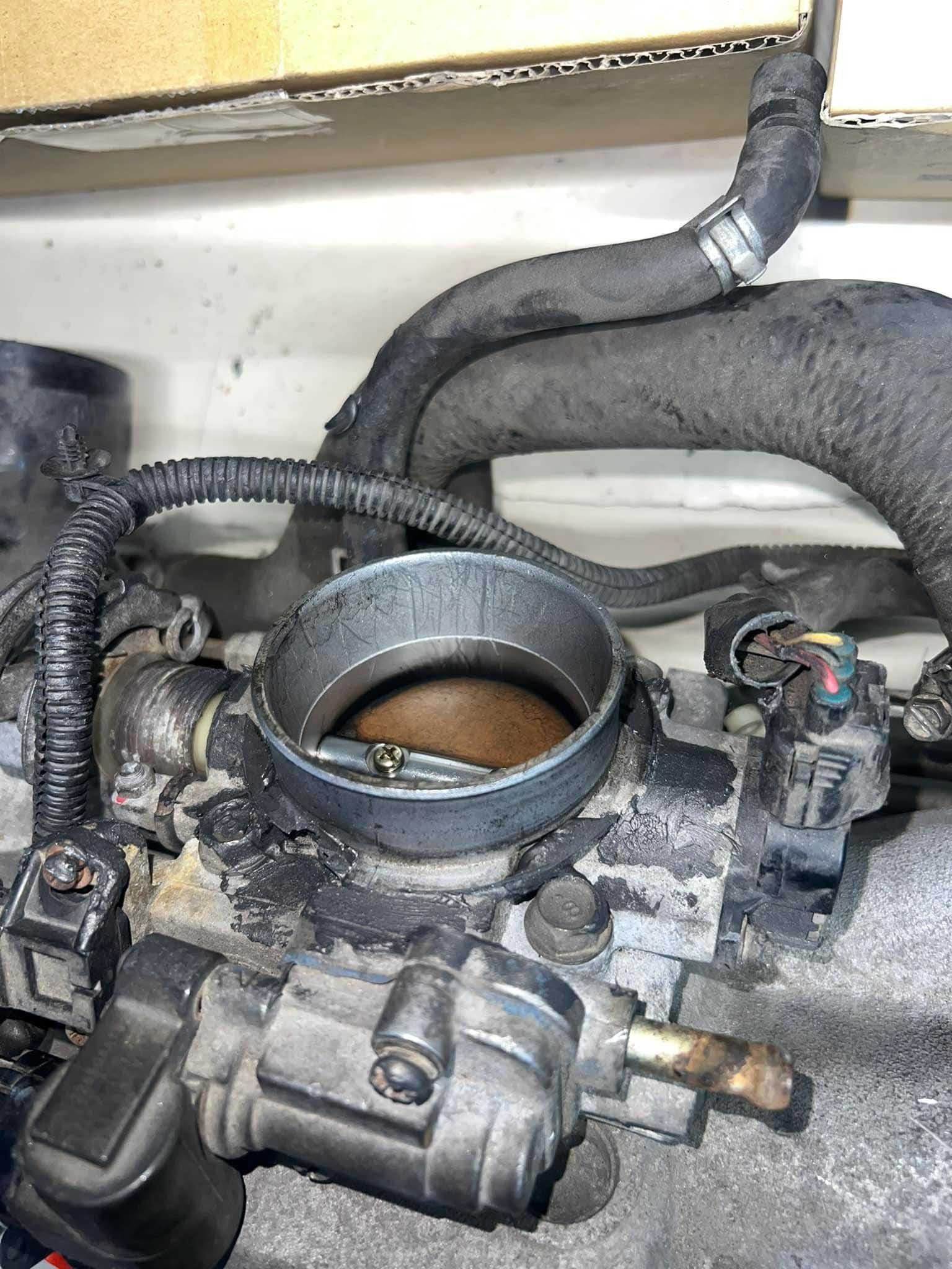 Engine throttle body with hoses and sensors, partially visible. Gray and black components, dusty.