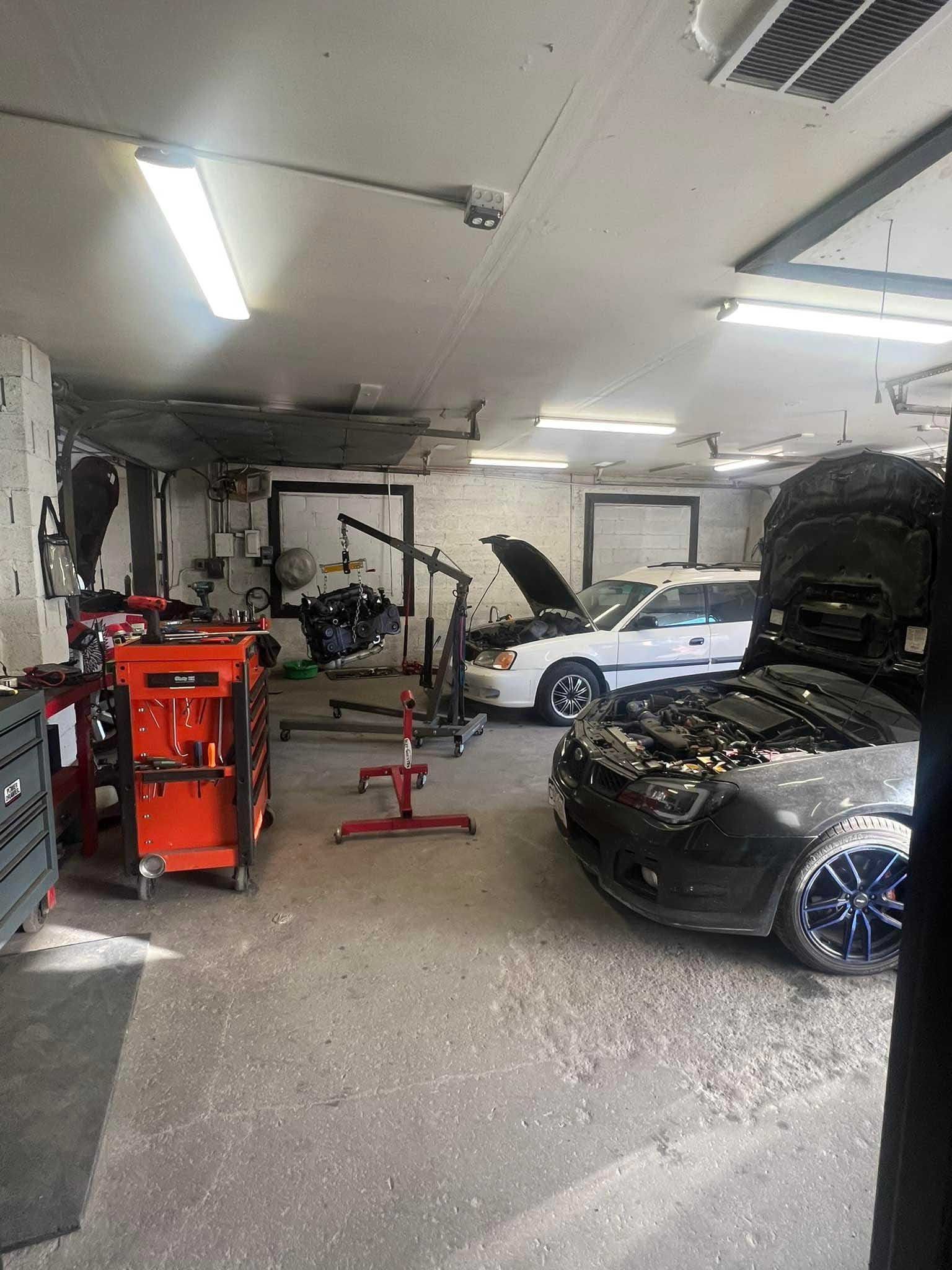 Cars in a cluttered auto repair shop, including a white sedan and black car with open hoods.