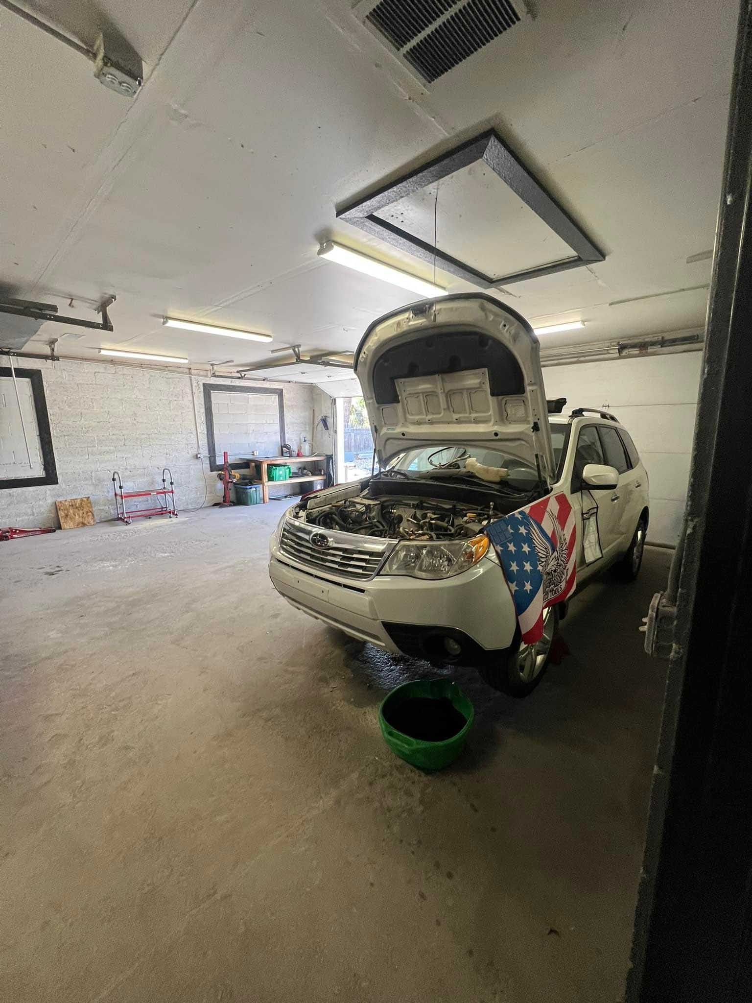 White SUV with open hood in a garage. Green oil pan on ground. American flag design on the side.