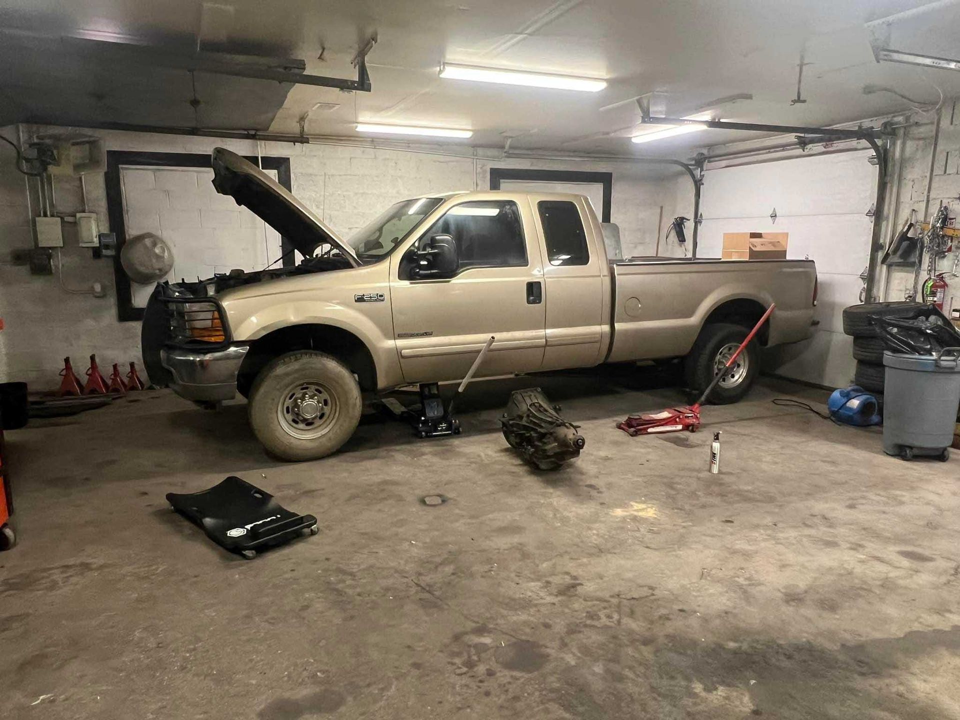 A tan pickup truck with hood up in a garage, a transmission is on the floor; tools scattered.