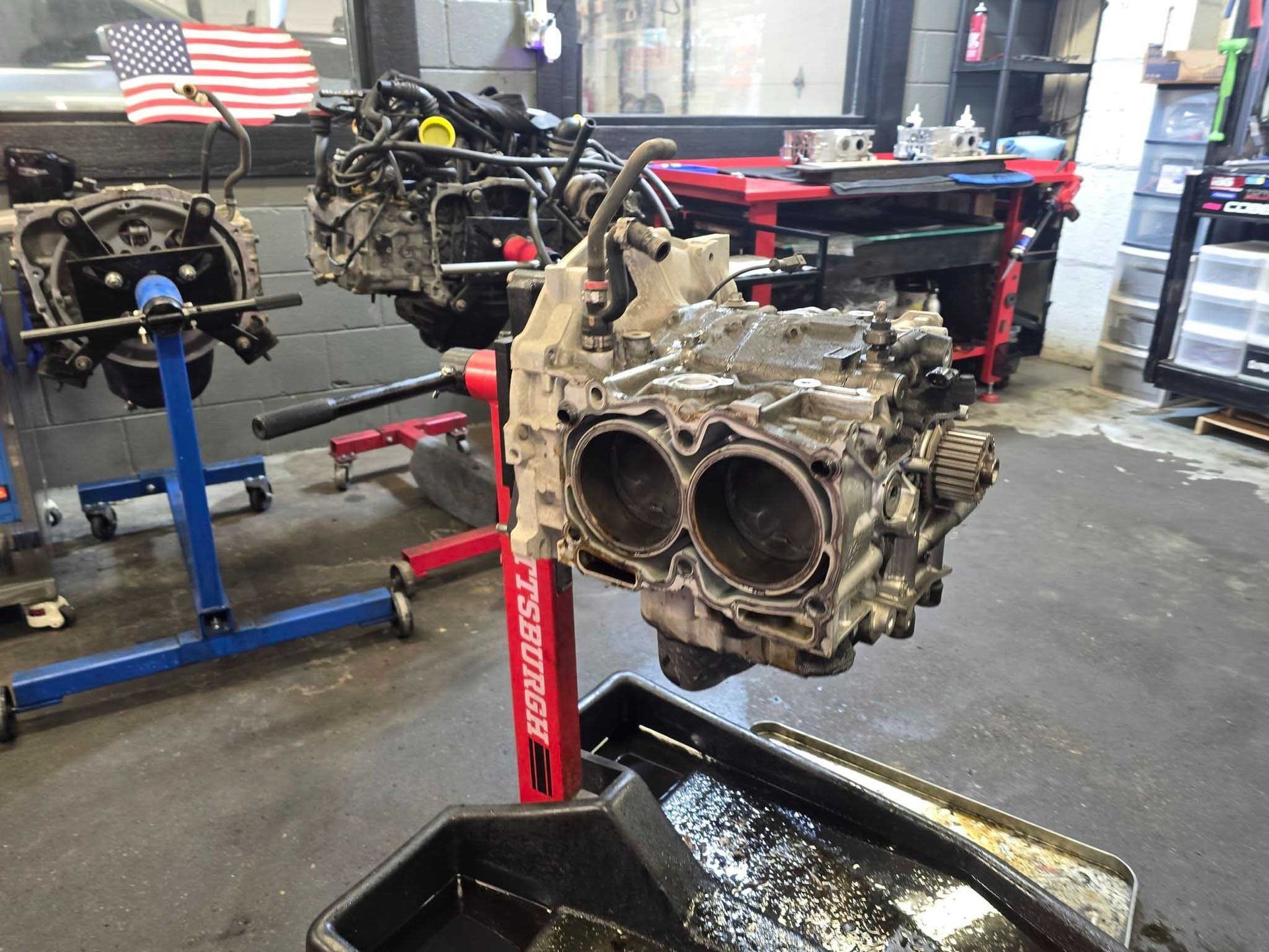 Car engine repair shop. Dismantled engine block on stand, oil pan below. Other engines and equipment visible.
