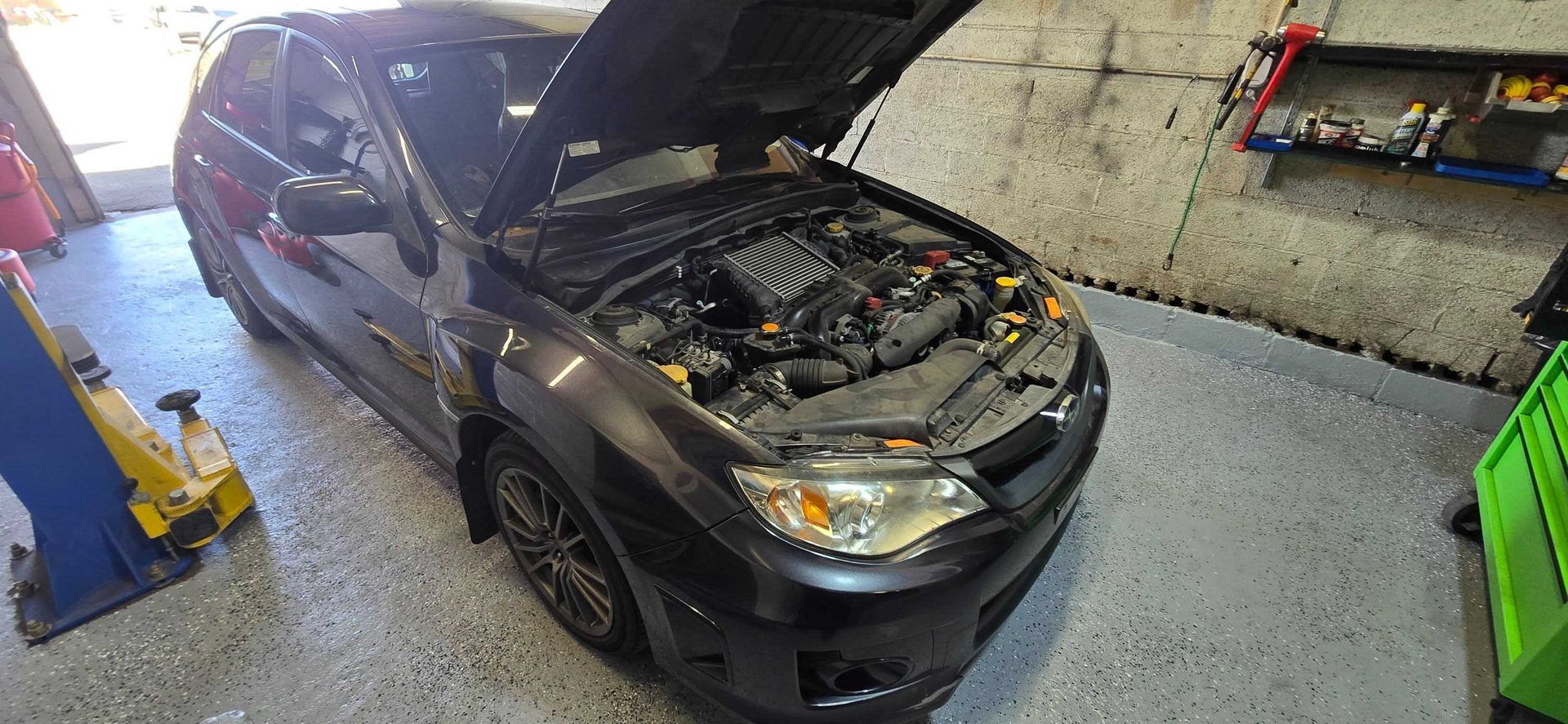Dark gray Subaru hatchback with hood open in a garage, possibly for repair.