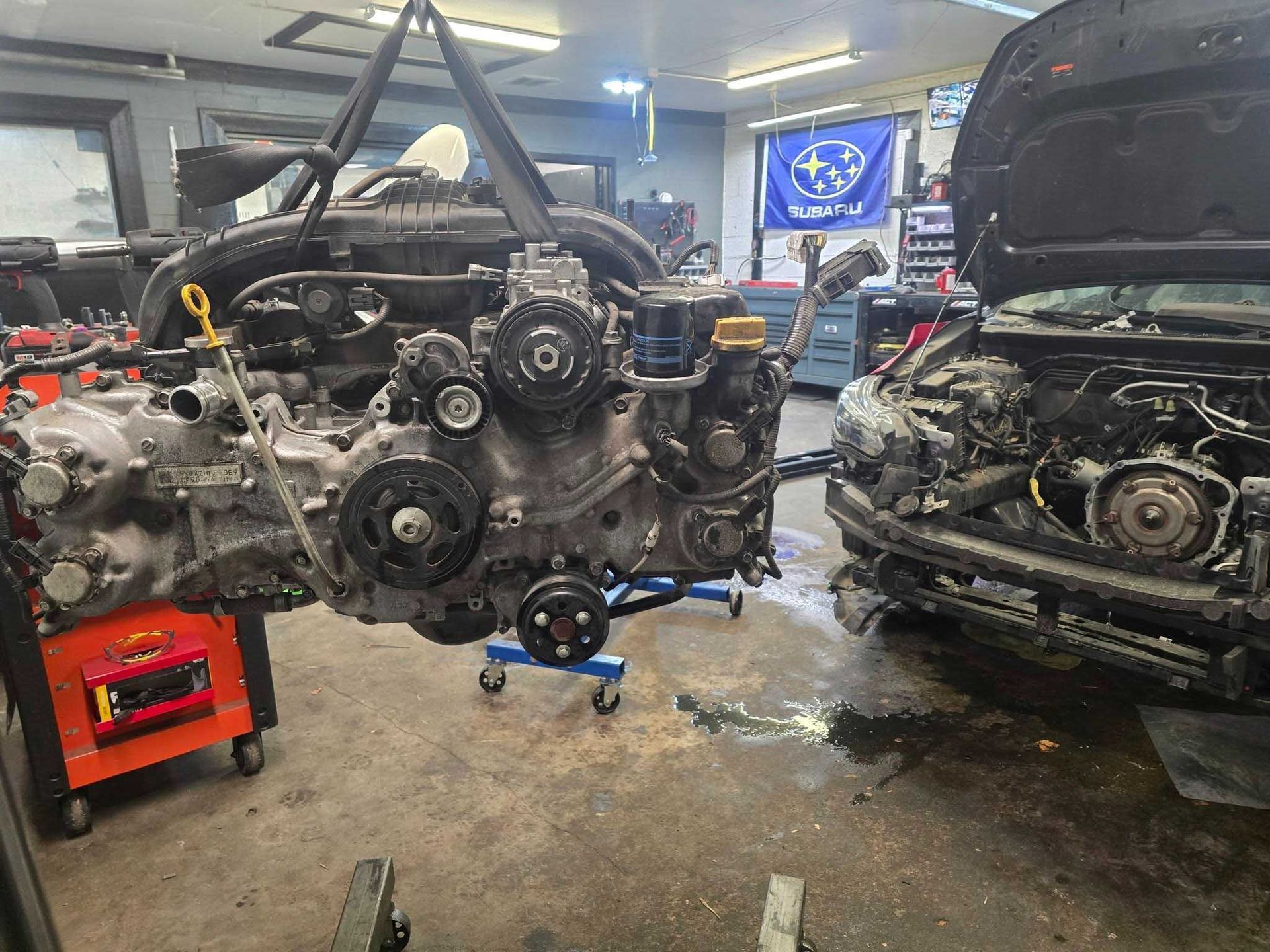 An engine suspended by a hoist in a Subaru auto shop, next to a car with an open hood.