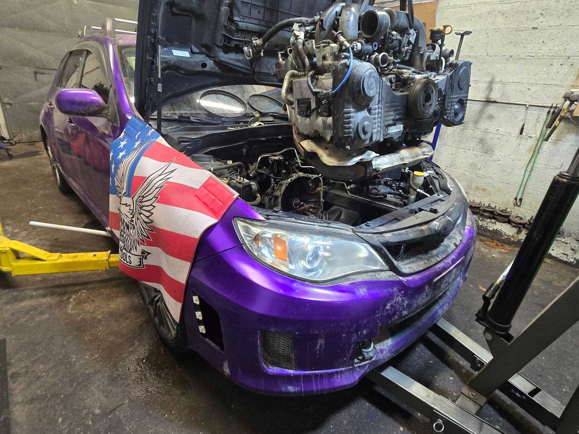 Purple Subaru with hood open, engine removed, American flag on front, in a garage.