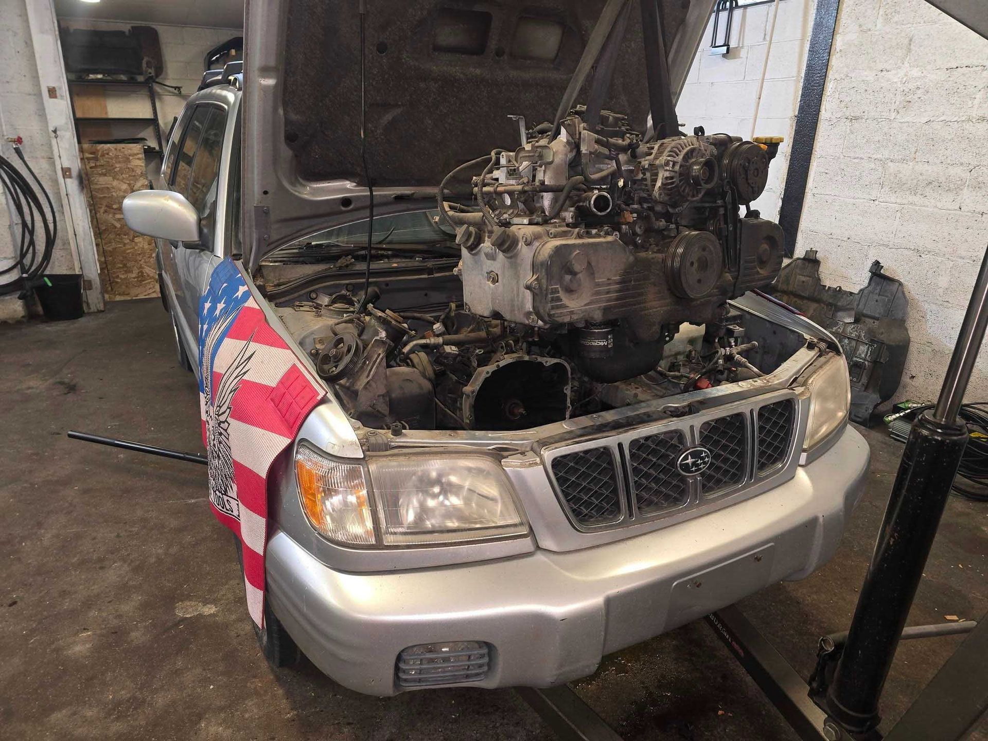 Silver Subaru Forester with open hood, engine removed, in a garage.