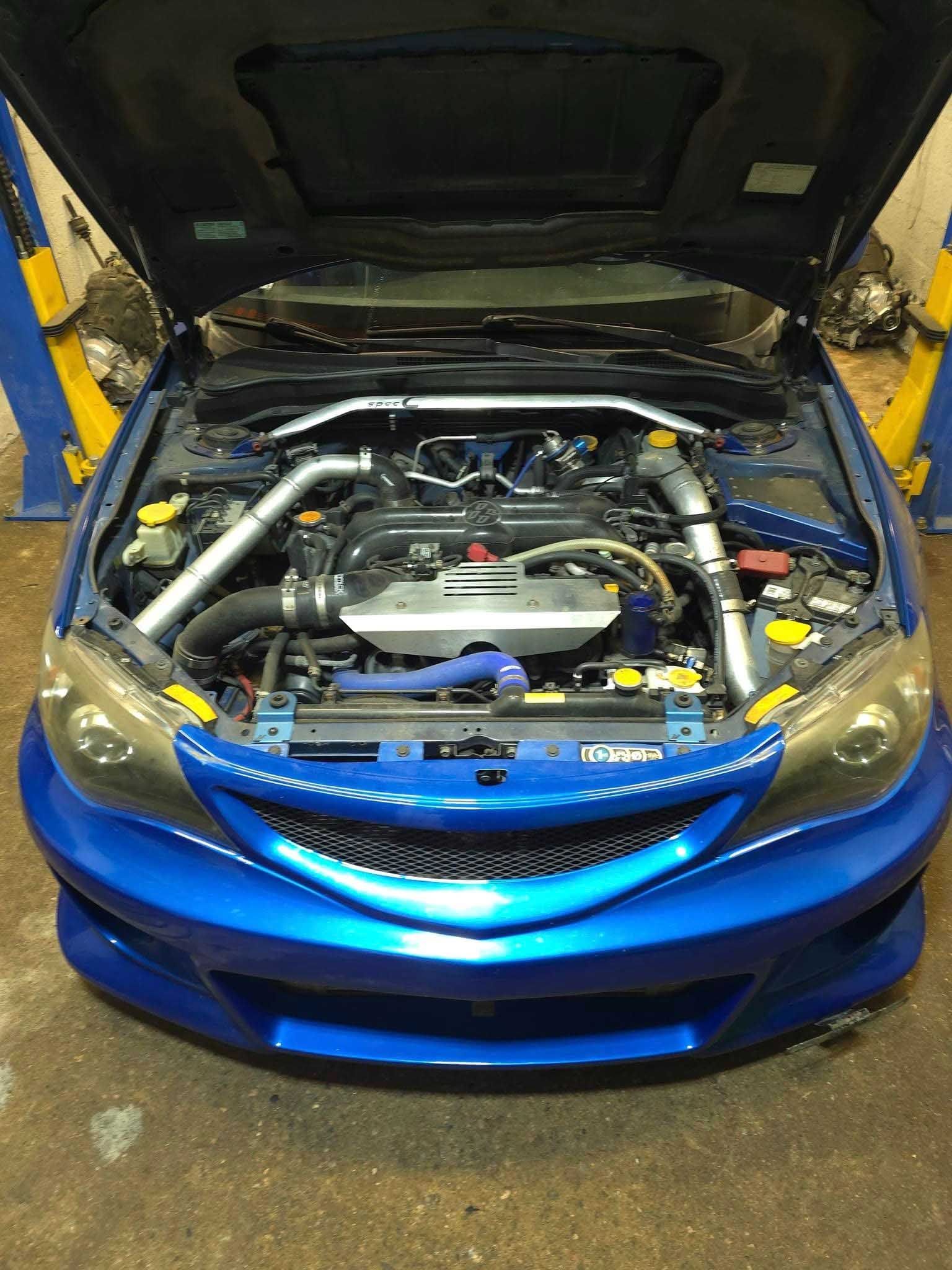 Blue Subaru Impreza with open hood, showing engine components in a garage.