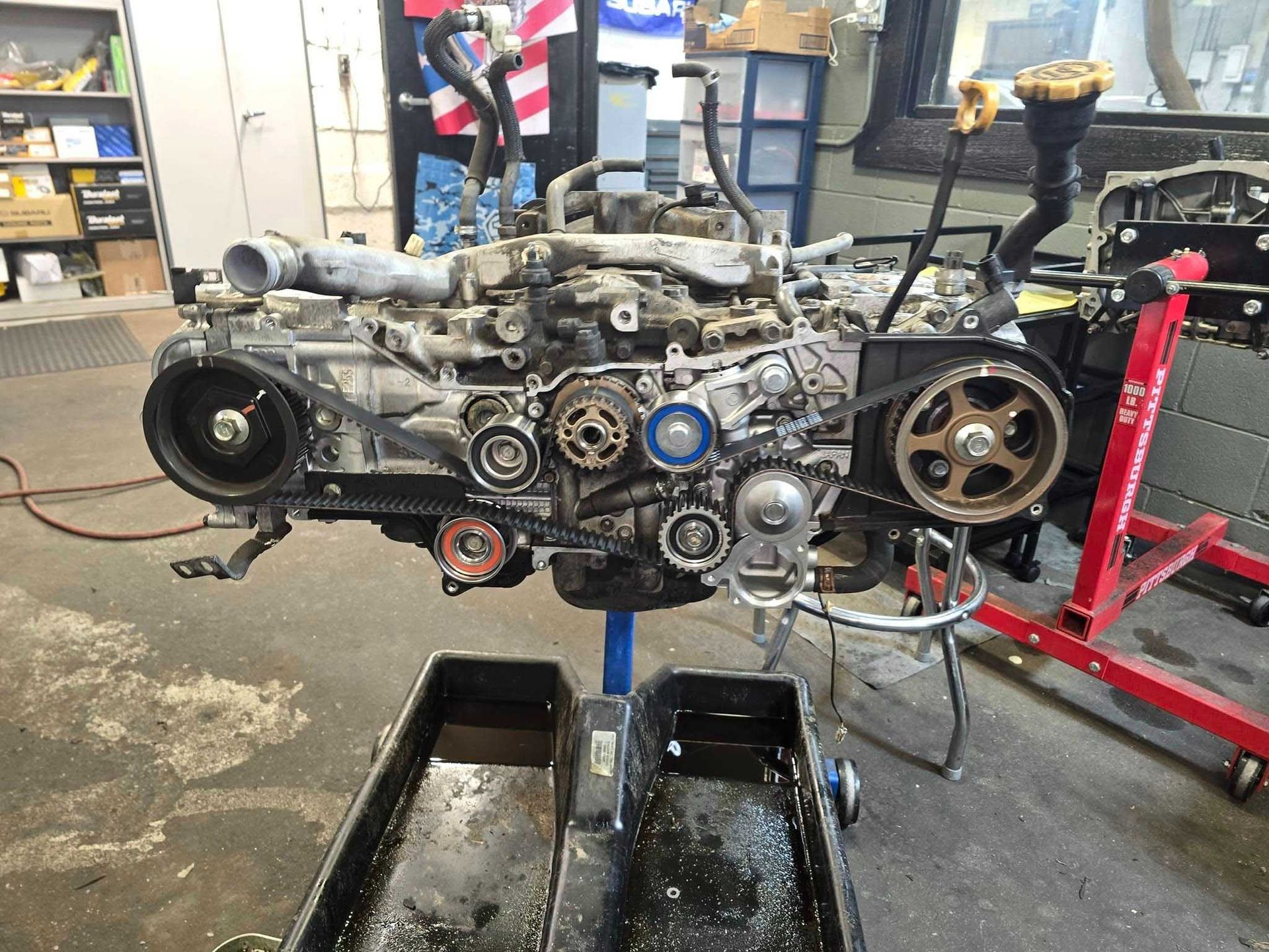 Engine on a stand with belt and pulley system, in a workshop setting. Oil pan underneath.