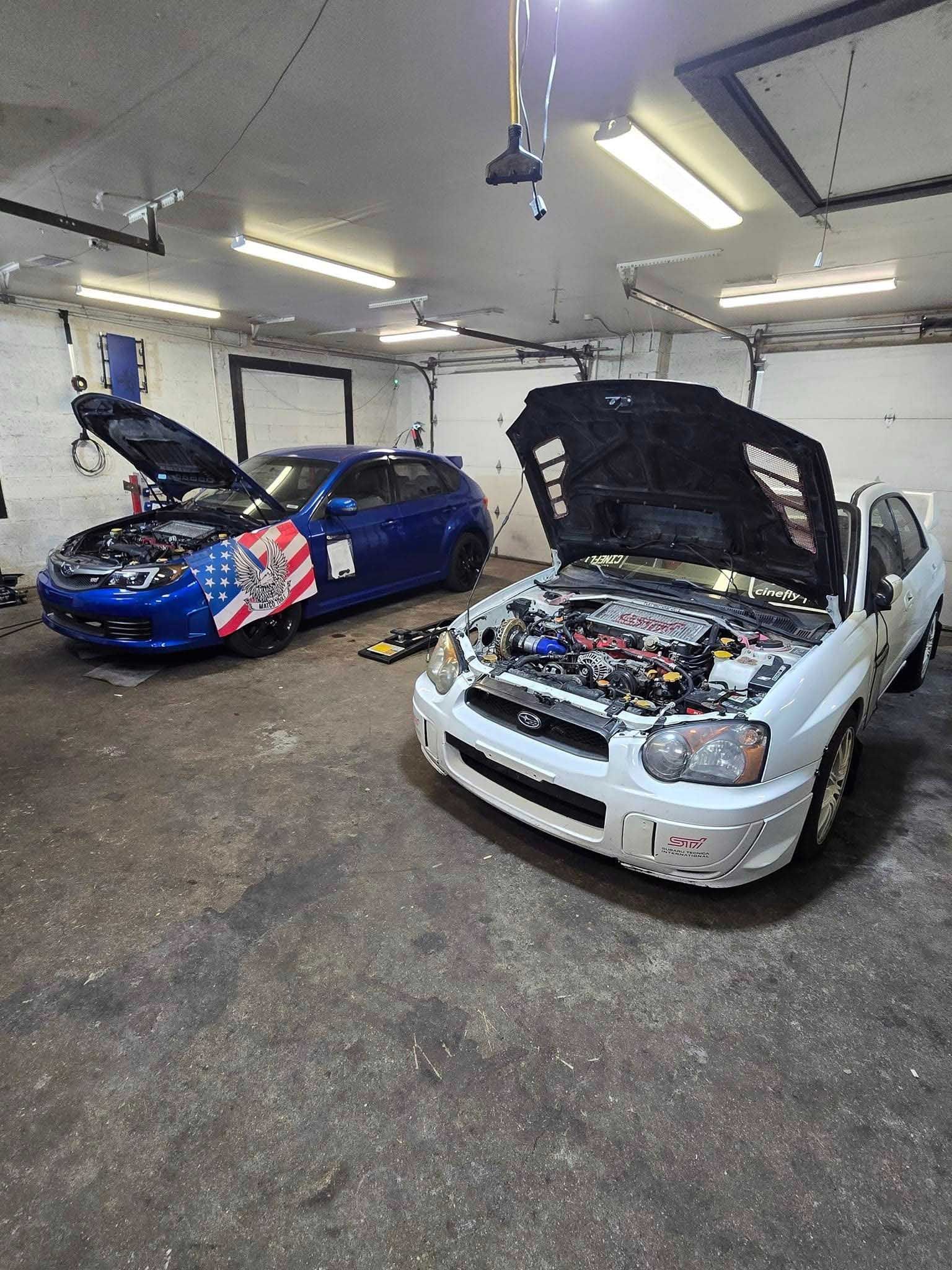 Two Subaru cars with open hoods in a garage. One is blue, the other is white.
